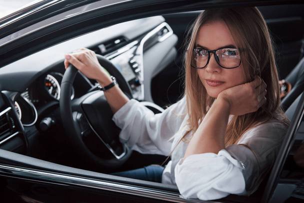 Great style. Beautiful blonde girl sitting in the new car with modern black interior. - Фото, изображение