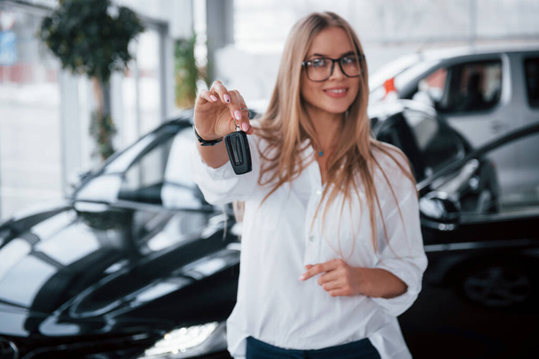 Holds keys by the fingers with pink nails. Young manager in the auto shop stands against perfectly polished black car. - Photo, Image