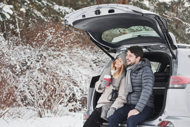 Smiling together. Nice couple have weekend at countryside at winter time. Sitting on the rear part of modern car. - Photo, image