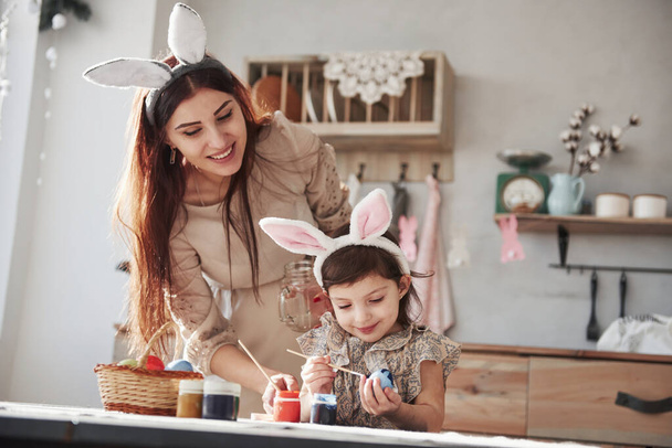 Painting egg in blue. Mother and daughter in bunny ears at easter time have some fun in the kitchen at daytime. - 写真・画像