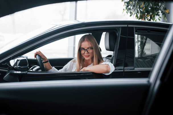 Expencive vehicle. Beautiful blonde girl sitting in the new car with modern black interior. - Valokuva, kuva