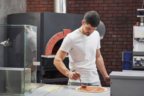 Stove is behind. Baker in white shirt putting sauce to make delicious pizza for an order in restaurant. - Foto, imagen