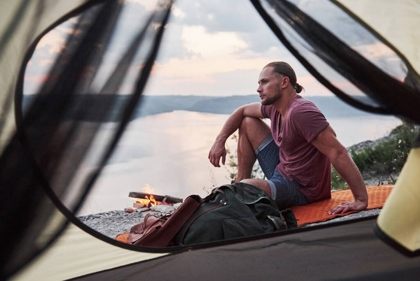 View from tent of traveller with backpack sitting on top of mountain enjoying view coast a river or lake. Traveling along mountains and coast, freedom and active lifestyle concept. - Foto, Imagen