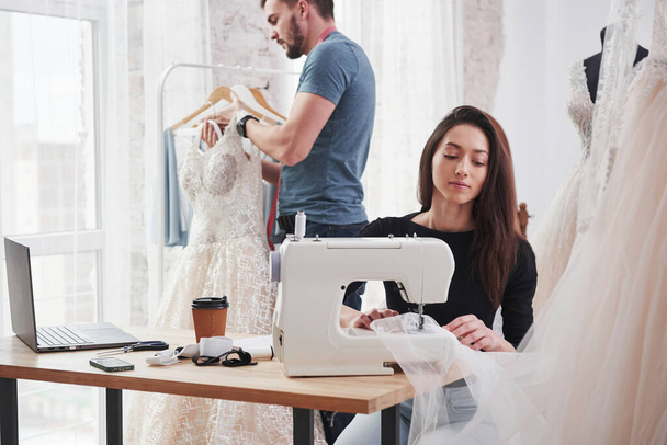 Guy is holding dress behind. Female fashion designer works on the new clothes in the workshop with man behind. - Foto, imagen