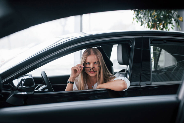 From the other car. Beautiful blonde girl sitting in the new car with modern black interior. - Fotó, kép