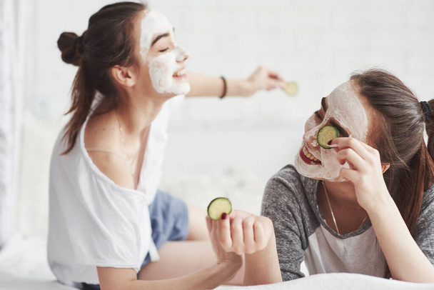 How this works. Conception of skin care by using fresh cucumber rings and white mask on the face. Two female sisters have weekend at bedroom. - Foto, immagini