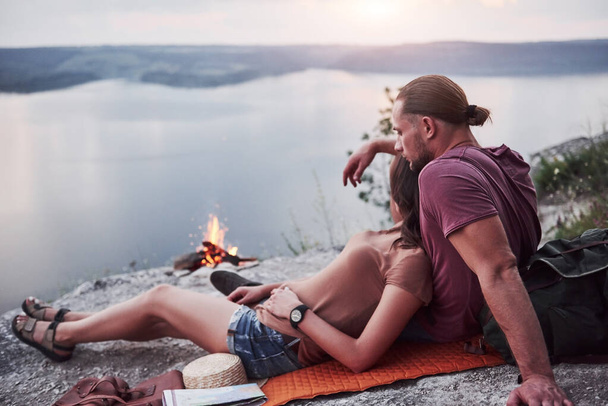 Hugging couple with backpack sitting near the fire on top of mountain enjoying view coast a river or lake. Traveling along mountains and coast, freedom and active lifestyle concept. - Foto, Imagen