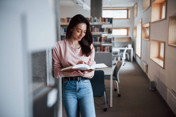 Standing and reading. Brunette girl in casual clothes having good time in the library full of books. - Foto, Bild