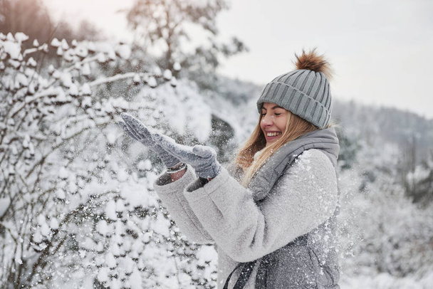 Playful mood. Cheerful girl in warm clothes throwing up snow outdoors near the beautiful forest. - Zdjęcie, obraz