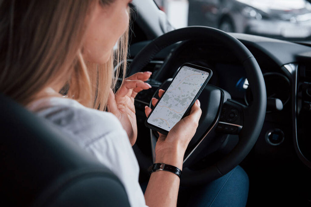 Mobile phone as tracker. Beautiful blonde girl sitting in the new car with modern black interior. - Foto, Imagem
