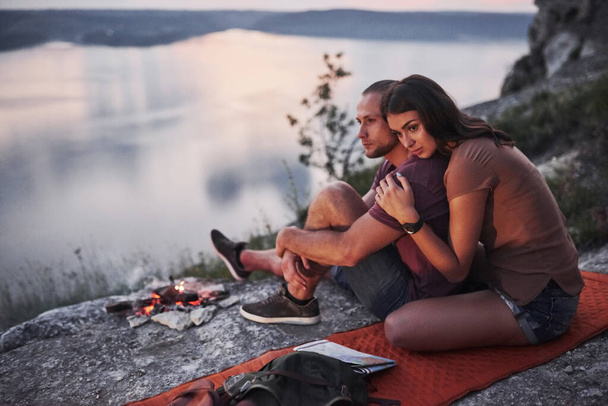 Hugging couple with backpack sitting near the fire on top of mountain enjoying view coast a river or lake. Traveling along mountains and coast, freedom and active lifestyle concept. - Fotografie, Obrázek