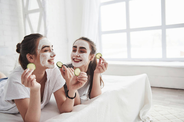 Going to eat that. Conception of skin care by using white mask and cucumbers on the face. Two female sisters have weekend at bedroom. - Photo, image