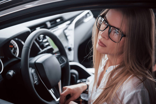 Dreamy look. Beautiful blonde girl sitting in the new car with modern black interior. - Foto, Imagen