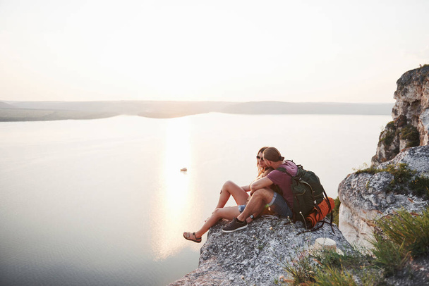 Hugging couple with backpack sitting on top of rock mountain enjoying view coast a river or lake. Traveling mountains and coast, freedom and active lifestyle concept. - Fotografie, Obrázek