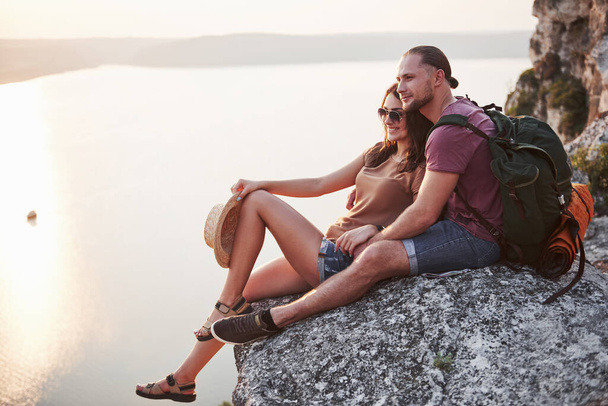 Hugging couple with backpack sitting on top of rock mountain enjoying view coast a river or lake. Traveling mountains and coast, freedom and active lifestyle concept. - Fotoğraf, Görsel