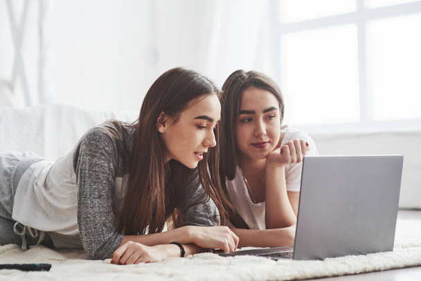 Sosyal medyada sörf yapmak. Genç bayan ikizler beyaz yatağın yanında yerde yatıyorlar ve gümüş renkli dizüstü bilgisayar kullanıyorlar.. - Fotoğraf, Görsel