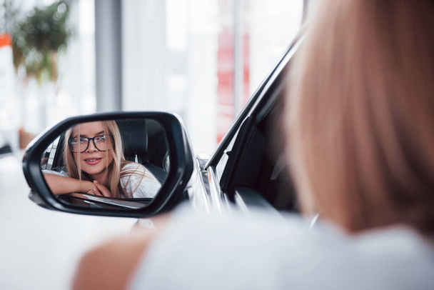 Ze zrcadla. Žena v brýlích dívá do bočního zrcátka moderního vozidla v salónu. - Fotografie, Obrázek