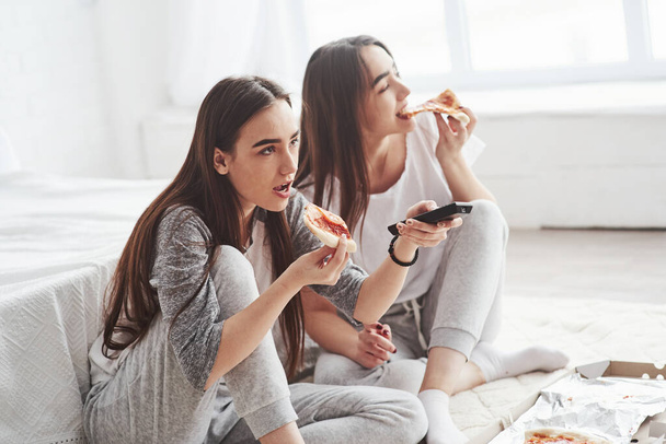 Nice looking young girls. Sisters eating pizza when watching TV while sits on the floor of beautiful bedroom at daytime. - Foto, Bild