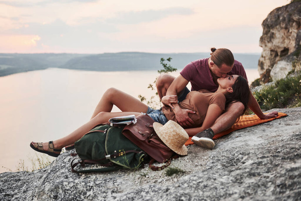 Hugging couple with backpack sitting on top of rock mountain enjoying view coast a river or lake. Traveling mountains and coast, freedom and active lifestyle concept. - Foto, afbeelding
