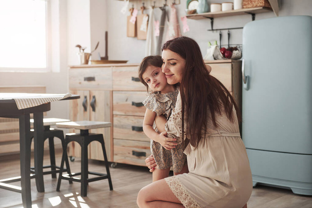 Mother sitting and hugs her daughter at the kitchen at daytime. - Foto, imagen