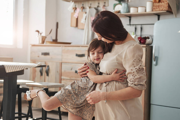 Sıcak atmosfer. Hamile kadın ayakta duruyor ve gündüz vakti mutfakta kızına sarılıyor.. - Fotoğraf, Görsel