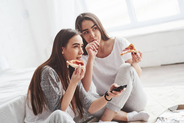 Girl licks her finger, it's so taste. Sisters eating pizza when watching TV while sits on the floor of beautiful bedroom at daytime. - Foto, imagen