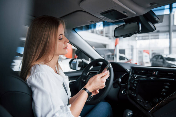 Checks the time. Beautiful blonde girl sitting in the new car with modern black interior. - Fotoğraf, Görsel