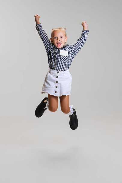 Full-length side view portrait of little cute girl, child in skirt and checkered shirt jumping isolated over gray background - Photo, Image