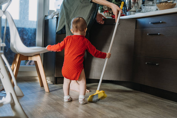 Little mothers helper. Cute toddler baby girl with mop help her mom do housework in kitchen. Cute little baby girl play with mop and makes cleaning the house - Photo, image
