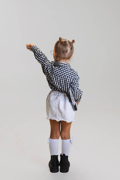 Full-length portret van klein meisje, kind in rok en geruit shirt wijzend ergens geïsoleerd over grijze achtergrond - Foto, afbeelding