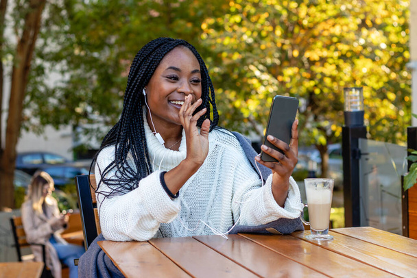 Egy gyönyörű fiatal afro-amerikai lány és egy szőke a háttérben, pihennek egy kávézóban és beszélgetnek a közösségi oldalakon a mobiljukon. Szabadidő és városi életmód. - Fotó, kép