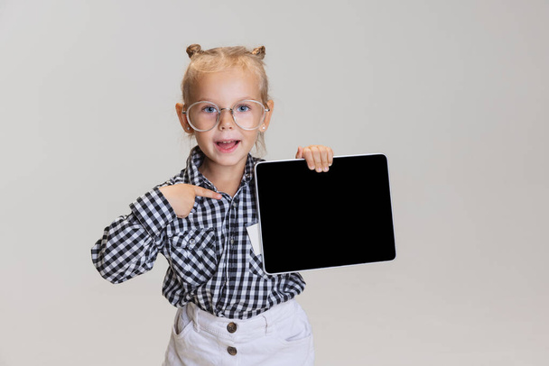 Cropped portrait of cute little girl, child pointing at tablet isolated over gray background. Boss kid - Фото, изображение