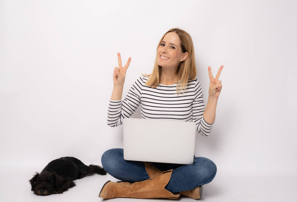 Фото молодой женщины, сидящей на полу с ноутбуком и маленькой собакой компаньон с табличкой победы изолированный белый фон - Фото, изображение
