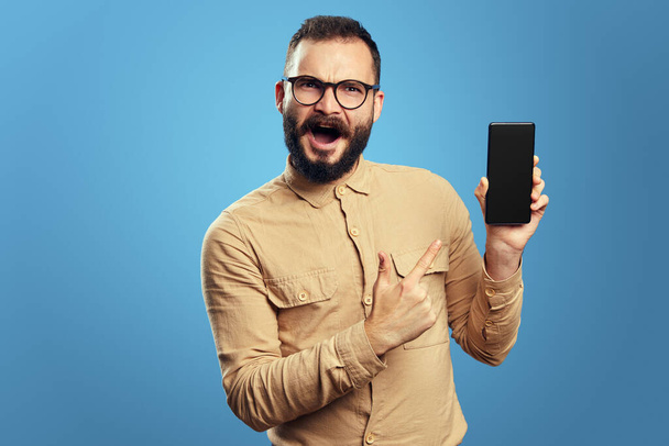 Junger Mann zeigt leeres Bildschirm-Handy, isoliert über blauem Hintergrund - Foto, Bild