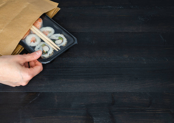 Χέρι λαμβάνοντας δίσκο σούσι από χάρτινη σακούλα σε μαύρο ξύλινο φόντο. Αντιγραφή χώρου. Έννοια τροφίμων. - Φωτογραφία, εικόνα