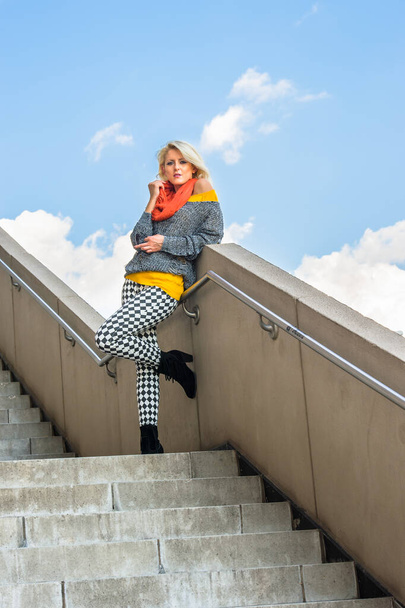 Dressing in a yellow underclothes, a gray fashionable sweater, black and white pattern pants, a red scarf, holding sunglasses, a young blonde girl is standing by railings of a stair , charmingly looking at you - Photo, image