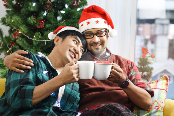 Adorable smiling young LGBT couple sharing special moment together on Christmas holiday, Asian gay male lover sitting on sofa, drinking hot beverage chocolate to celebrate night before Christmas. - Photo, Image