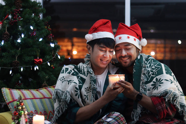 Adorável sorrindo jovem casal LGBT compartilhando um cobertor e tendo momento quente especial juntos no feriado de Natal, amante gay asiático sentado no sofá na sala de estar para celebrar a noite de Natal.   - Foto, Imagem
