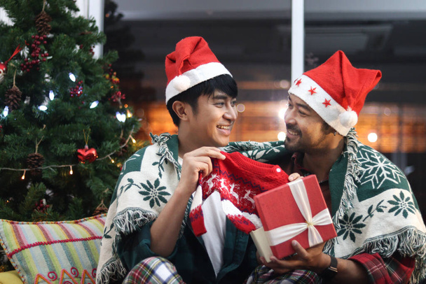 Adorable smiling young LGBT couple celebrate Christmas night holiday, open gift box, hold kid sweater for their adopted child. Two Asian gay lover plan to build a family, create future together. - Photo, Image