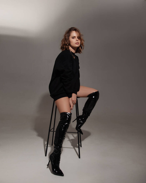 beautiful woman posing on white studio background. model wearing black sweatshirt and black boots. lady sitting on bar chair. fashion portrait - Fotografie, Obrázek