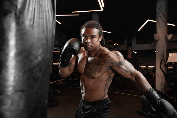 Afrikalı bir Amerikalı spor salonunda antrenman yapıyor ve boks torbası yumruklarını yumrukluyor. Spor salonu, fitness, boks, başarı, egzersiz ve güç kavramı - Fotoğraf, Görsel