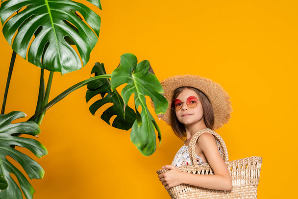 Cute little girl in straw hat in sunglasses holds straw handbag while stands near tropical plants - Фото, зображення