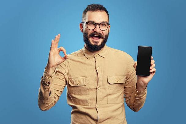 Молодий бородатий чоловік показує порожній екран мобільного телефону, жестикулюючи знак ОК
 - Фото, зображення