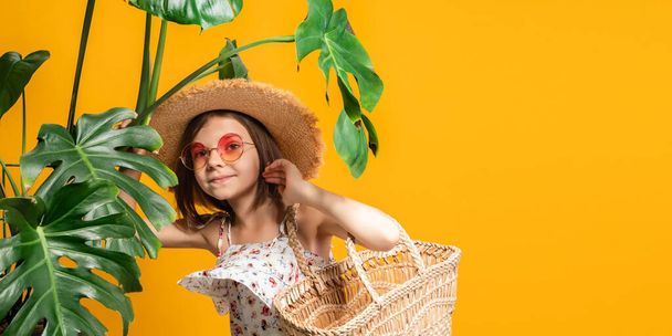 Cute little girl in straw hat, orange sunglasses holds straw handbag while stands in tropical plants - Photo, image