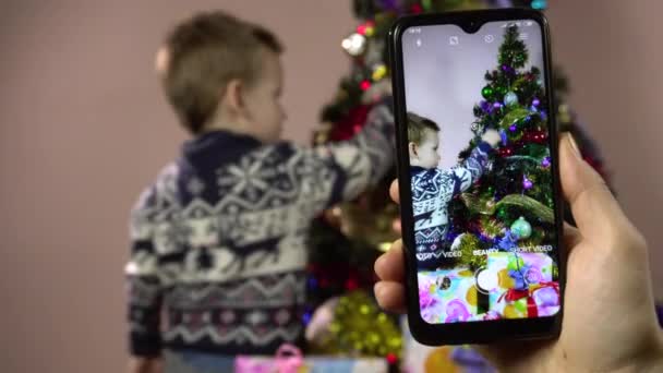 Küçük çocuk Noel ağacını süslüyor ve akıllı telefondan çekim yapmak için poz veriyor.. - Video, Çekim