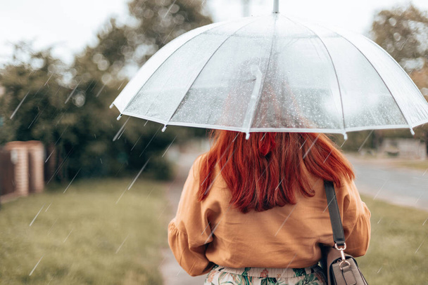 Zła pogoda. Widok z tyłu młodej kobiety w jasnych ubraniach pod przezroczystym parasolem w parku miejskim - Zdjęcie, obraz
