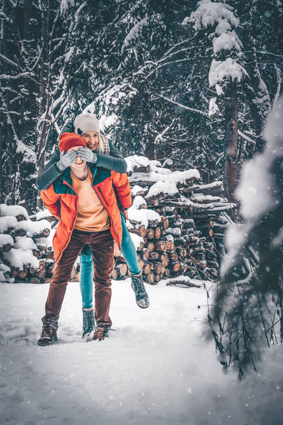 Gyönyörű, boldog fiatal férfi és nő bolondoznak egy havas téli erdőben. A nő hátulról ráakaszkodik a férfira, és eltakarja a szemét a kezével. Gyönyörű téli hangulatú fotó az emberekkel - Fotó, kép
