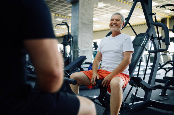 Elderly man poses on exercise machine in gym - Photo, image