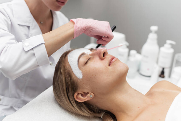 Beautician applies a white clay face mask with a brush on a womans face. Professional beautician applies white cosmetic cream to a girl lying on a couch in a modern spa. Close up. Relaxed woman - Photo, Image