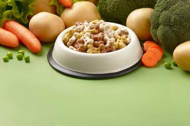 Dry dog vegan vegetarian food concept. Raw vegetables and green near bowl with pet feed on green background - Zdjęcie, obraz